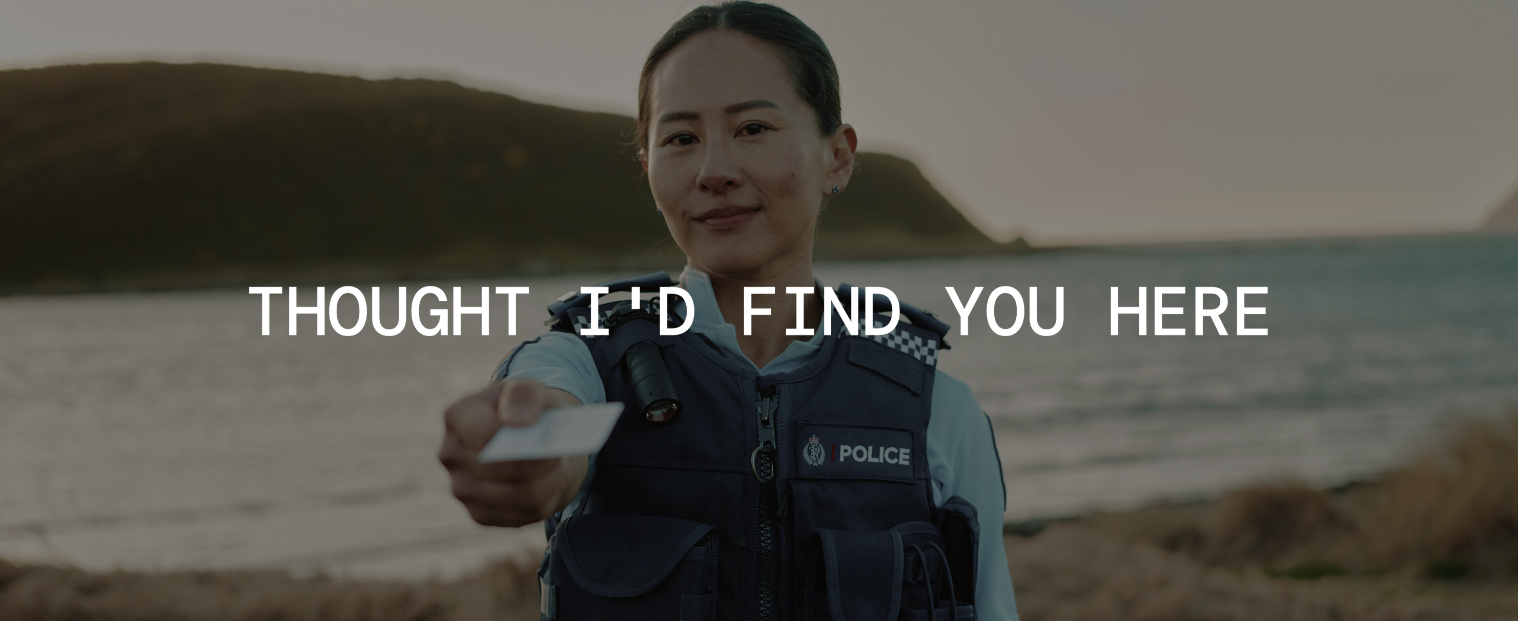 Police Officer standing on beach holding out a card towards the viewer. Text over image 'Thought I'd find you here